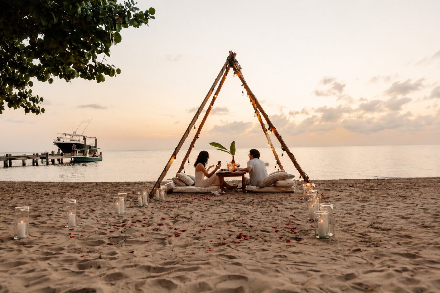 A candlelit table for two under a driftwood canopy on the sand at sunset